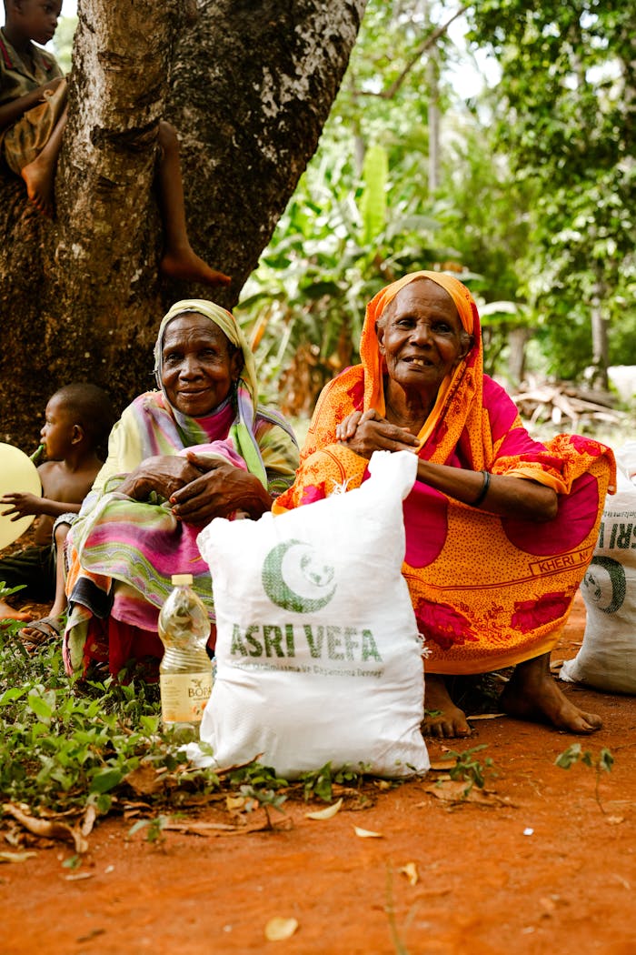 Two elderly women hold aid packages in a rural outdoor setting, sitting on red soil.