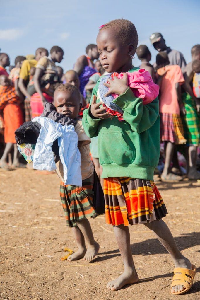 Two children carrying colorful clothes outdoors on a sunny day, surrounded by a group.